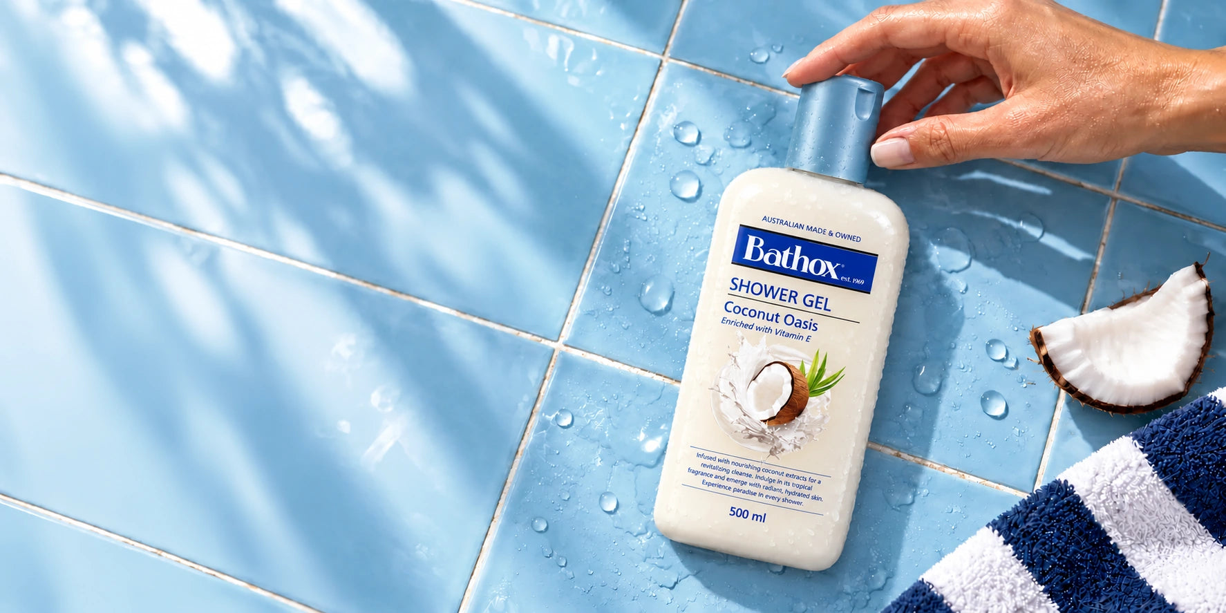 Coconut Oasis on wet blue tiles, with a hand resting on its cap and a striped towel.