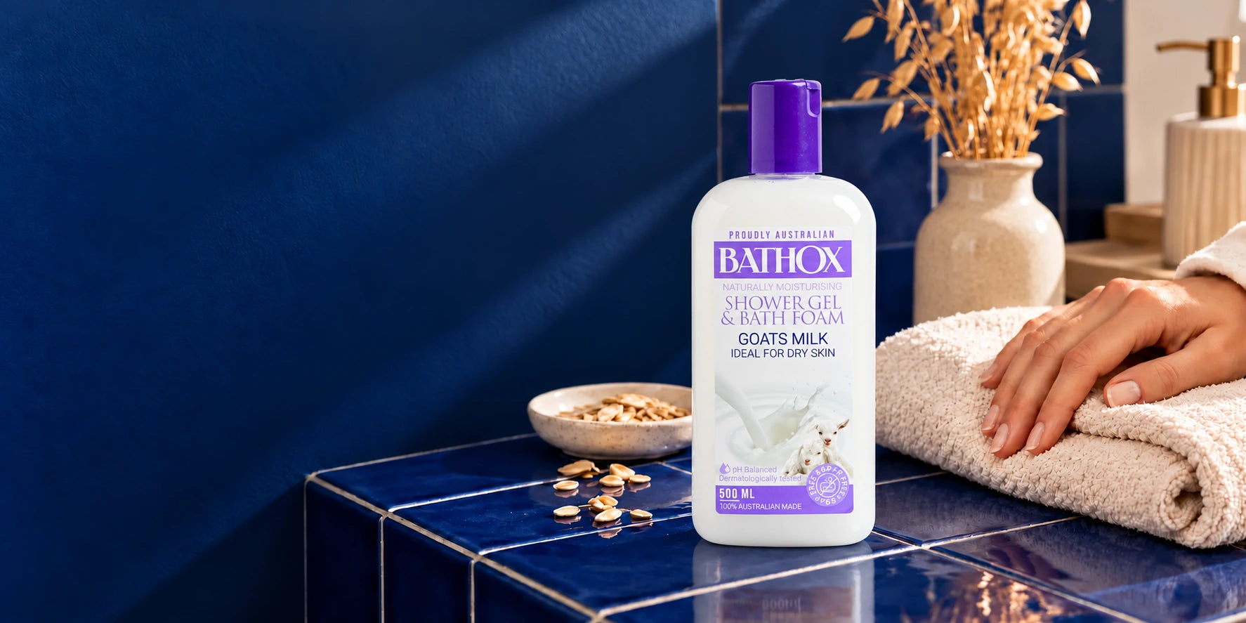 Goat’s Milk on deep blue tiles with a hand, a soft cotton towel and oat textures.
